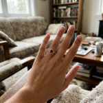 A candid, realistic photo of a hand displaying a ruby halo ring, with warm ambient lighting in a comfortable, everyday home environment showing a sofa and bookshelves.